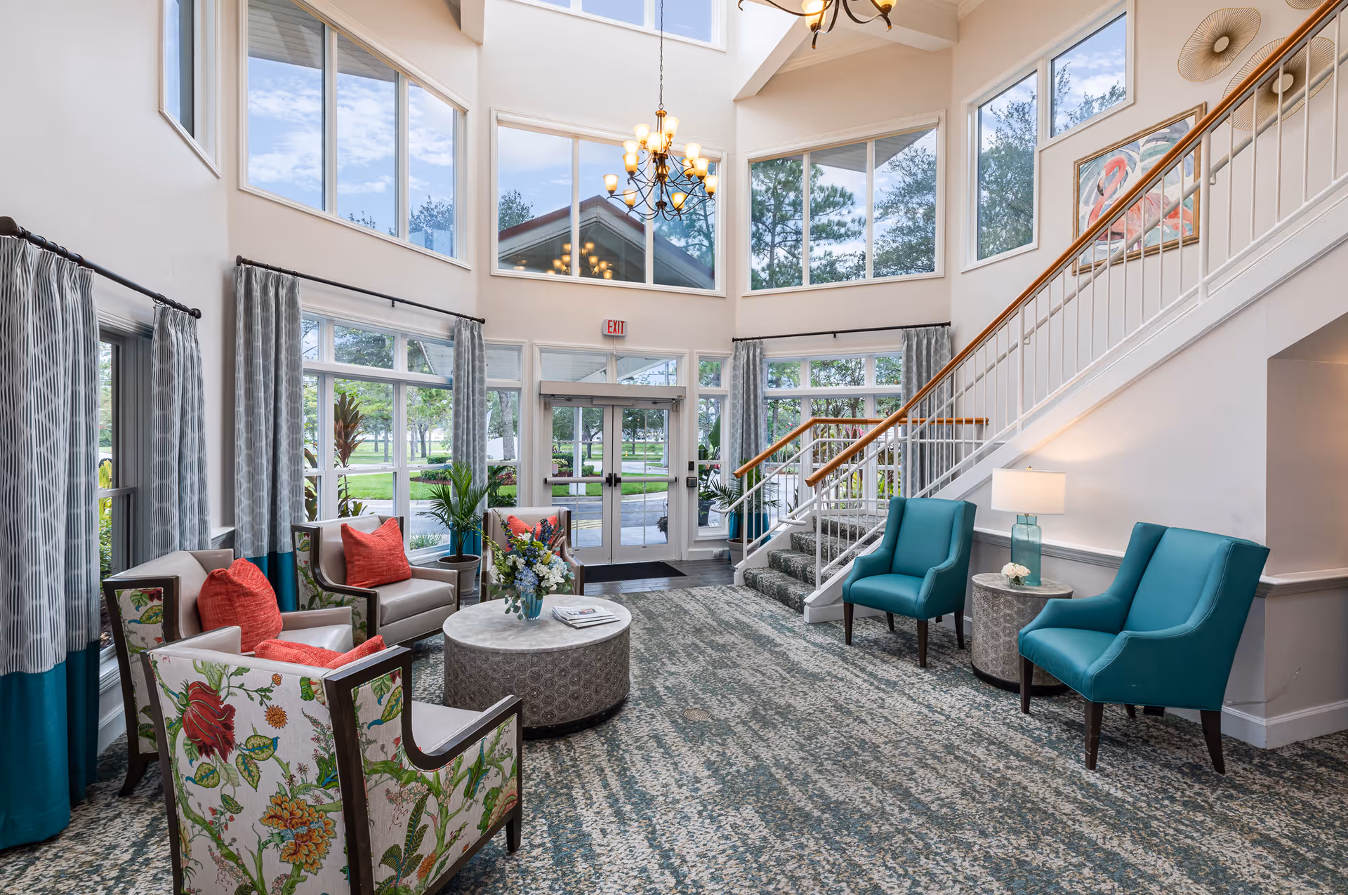 Bright and spacious senior living facility lobby with large windows, a high ceiling, and a chandelier. The room features a seating area with floral and teal armchairs, red accent pillows, a round coffee table with a flower arrangement, and a staircase leading to the upper floor. Outside greenery is visible through the windows.