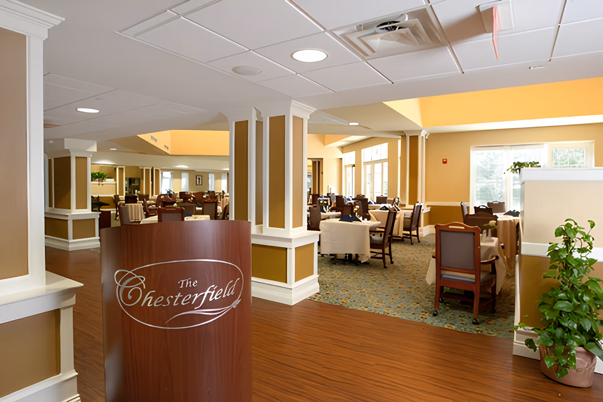 Interior view of a dining room area with a wooden welcome podium labeled 'The Chesterfield', multiple dining tables and chairs, and warm yellow-beige decor.