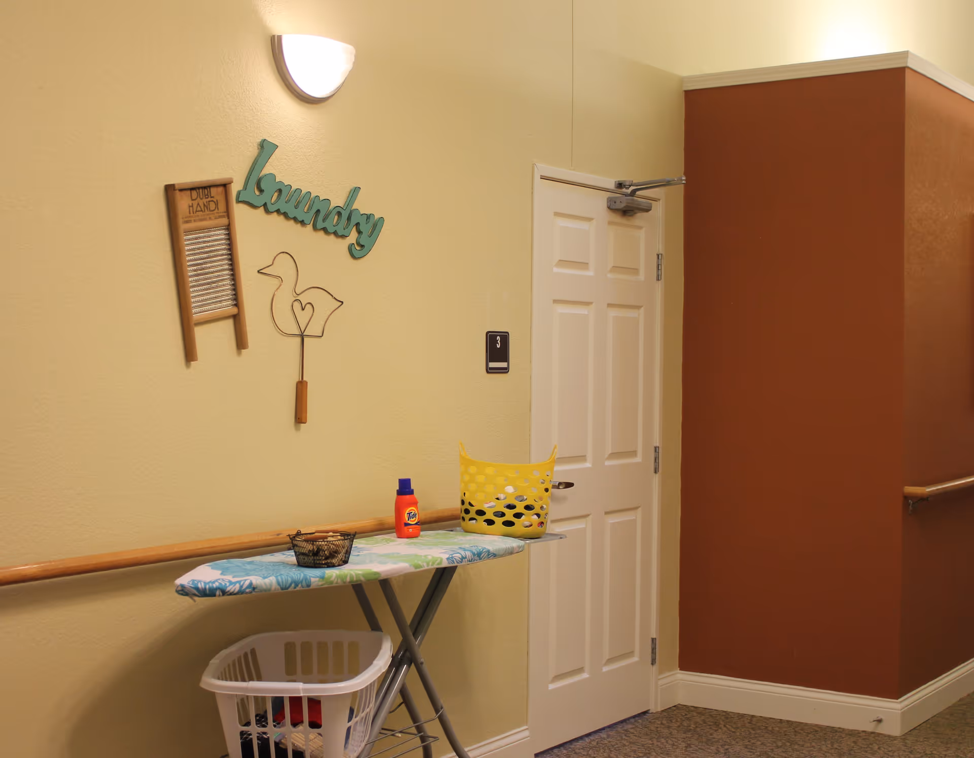 Interior hallway corner with a white door labeled number 3, a wall-mounted light fixture, and laundry-themed decorations including a washboard, a wire duck with a heart, and the word 'Laundry' in teal letters. An ironing board with a floral cover holds a yellow laundry basket and a bottle of Tide detergent, with a white laundry basket underneath containing clothes.