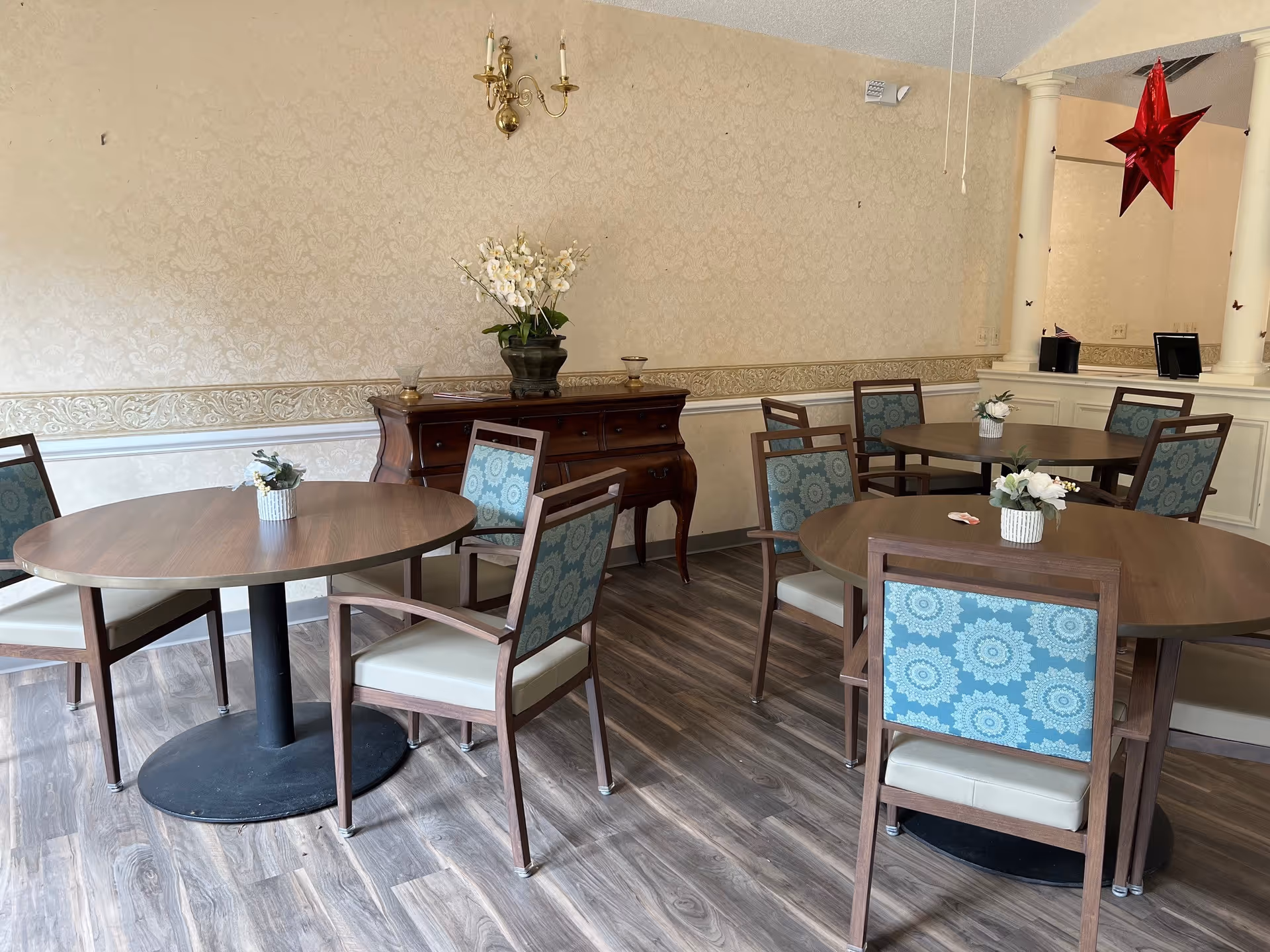 Interior view of a dining room with round wooden tables and chairs with blue patterned backs and beige seats. A wooden sideboard with a flower arrangement is against the wall, which has a beige patterned wallpaper. A red star decoration hangs from the ceiling near two white columns.