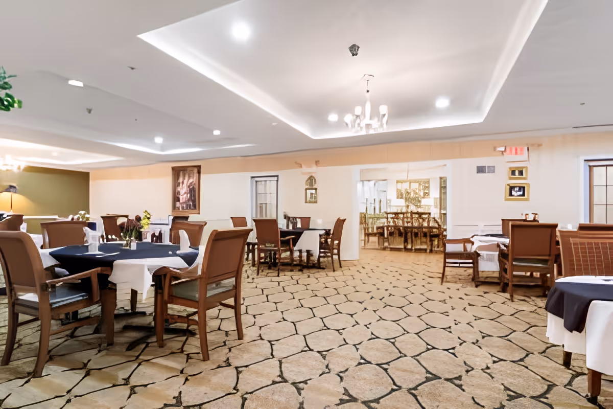 A spacious dining room with round and rectangular tables set with tablecloths and wooden chairs under recessed lighting and a chandelier.