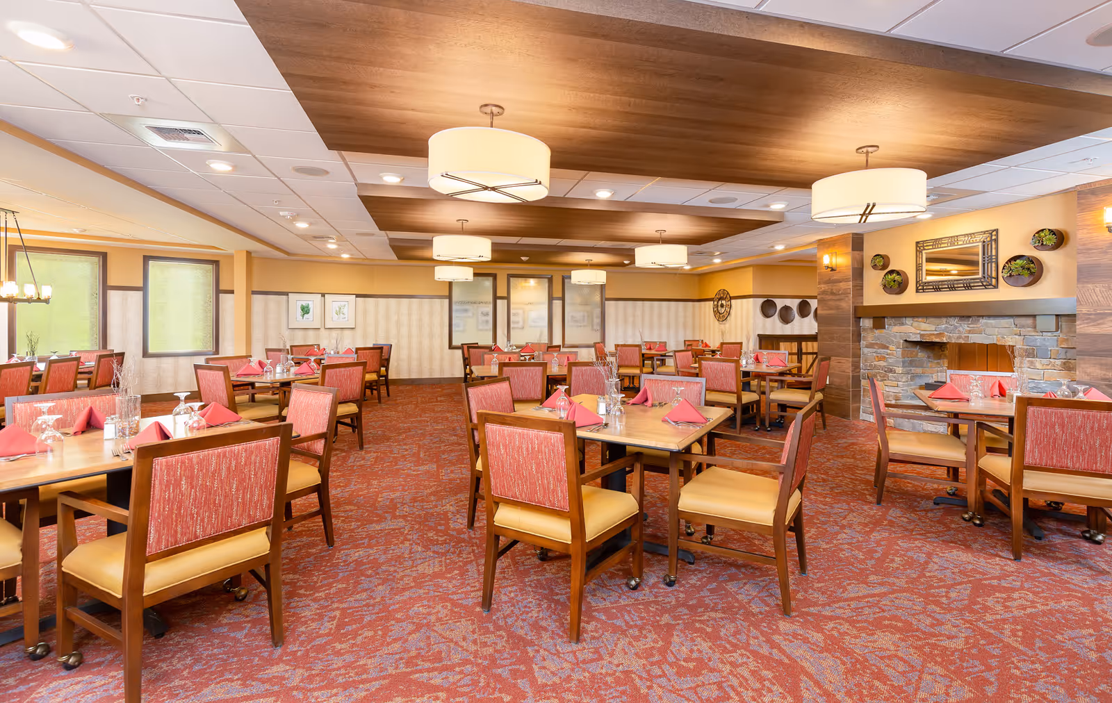 A spacious dining room with multiple wooden tables and chairs arranged neatly. Each table is set with red napkins, glasses, and silverware. The room features a patterned red carpet, warm yellow walls, decorative wall art, and a stone fireplace. Ceiling lights with round lampshades provide soft lighting.
