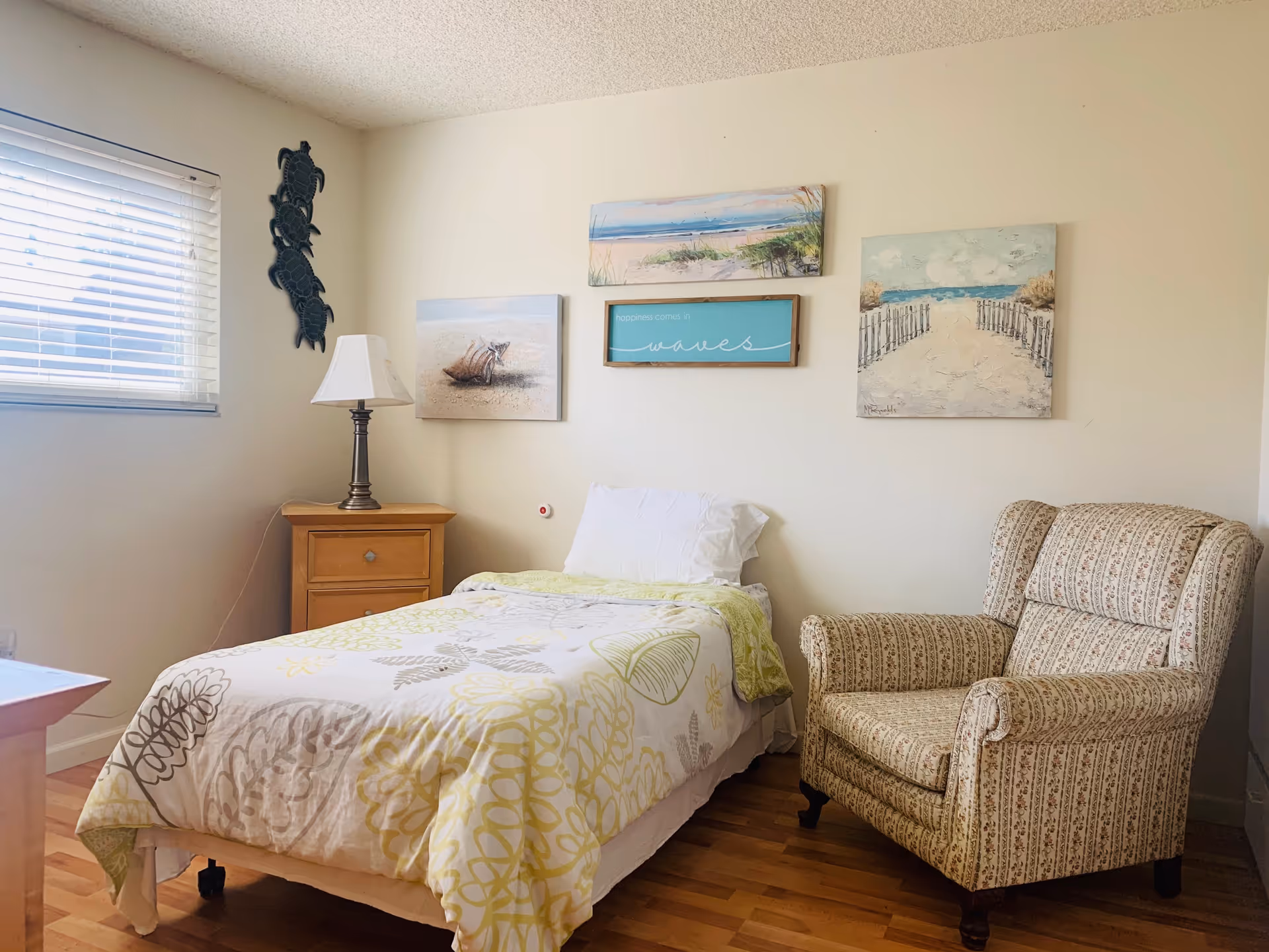 A bright single bedroom with a twin bed, wooden nightstand and lamp, patterned armchair, and beach-themed wall art.