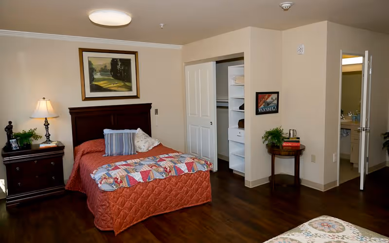 A cozy bedroom in a memory care community featuring a single bed with a red quilt and decorative pillows, a wooden nightstand with a lamp and small plant, an open closet with shelves and hanging space, a small round table with books and a plant, and a doorway leading to a bathroom.