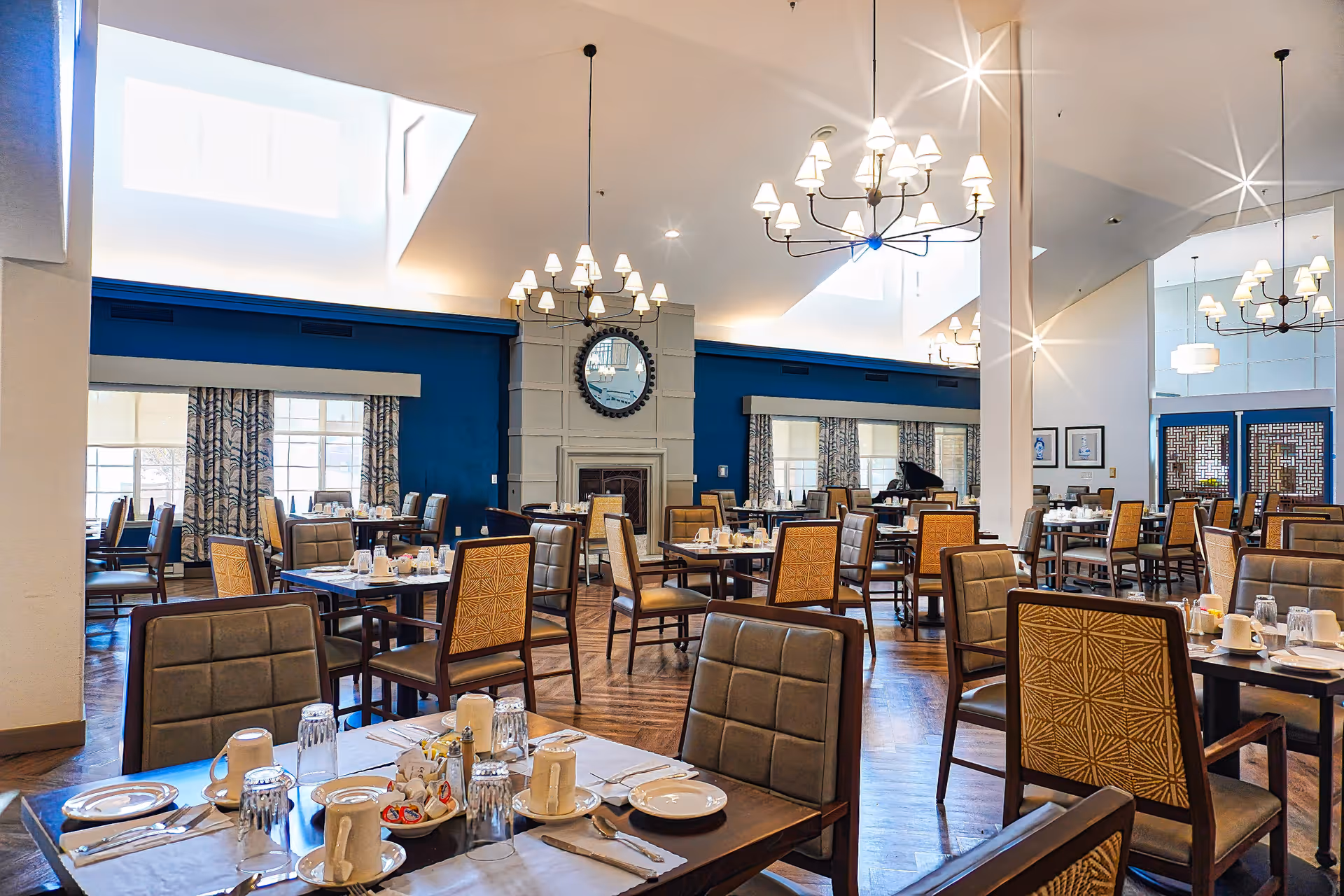 A spacious and well-lit dining room with multiple tables set with plates, cups, glasses, and silverware. The room features high ceilings with modern chandeliers, large windows with patterned curtains, a fireplace with a round mirror above it, and wooden flooring. The walls are painted blue and white, and there is a grand piano in the background.