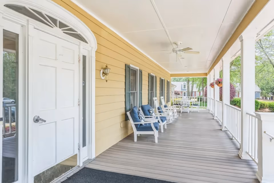 Covered front porch with a white entry door, several rocking chairs with blue cushions, a ceiling fan and white railing overlooking landscaped grounds.