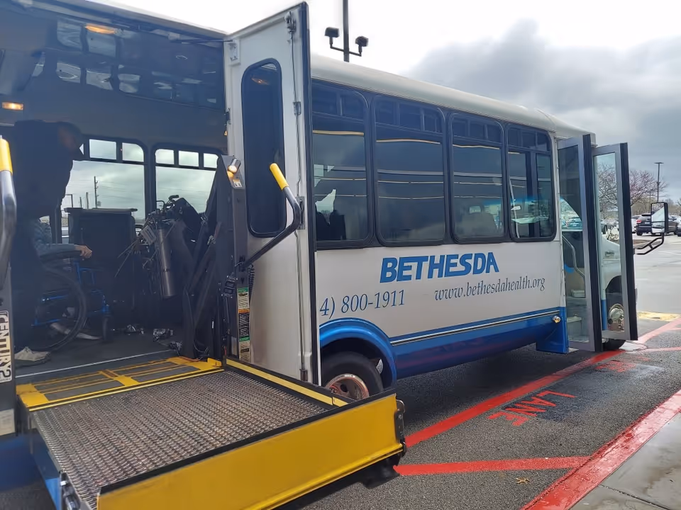 A Bethesda shuttle bus with an open wheelchair lift ramp at the side door. A person is assisting a wheelchair inside the bus. The bus is parked on a wet pavement with red painted markings.