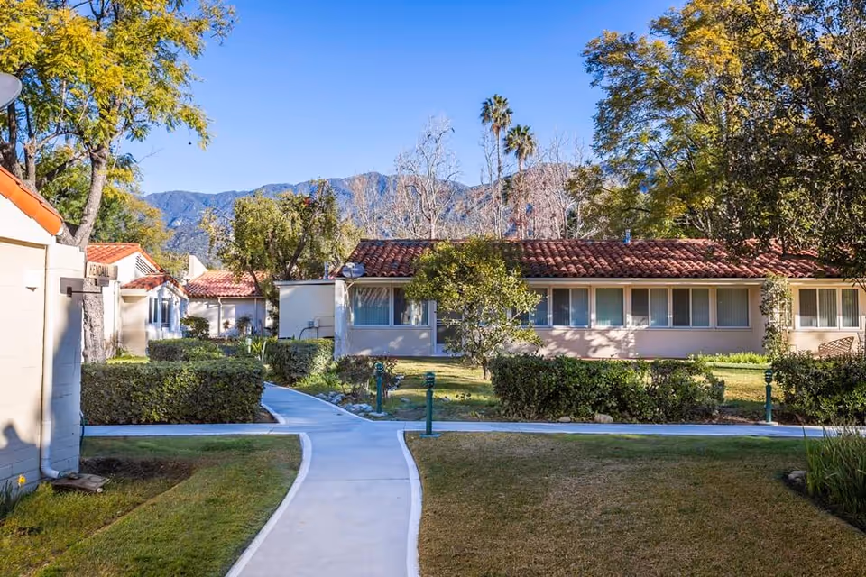 Walkway through a landscaped courtyard with single-story, red-tile-roofed buildings, trees, and mountains in the background.