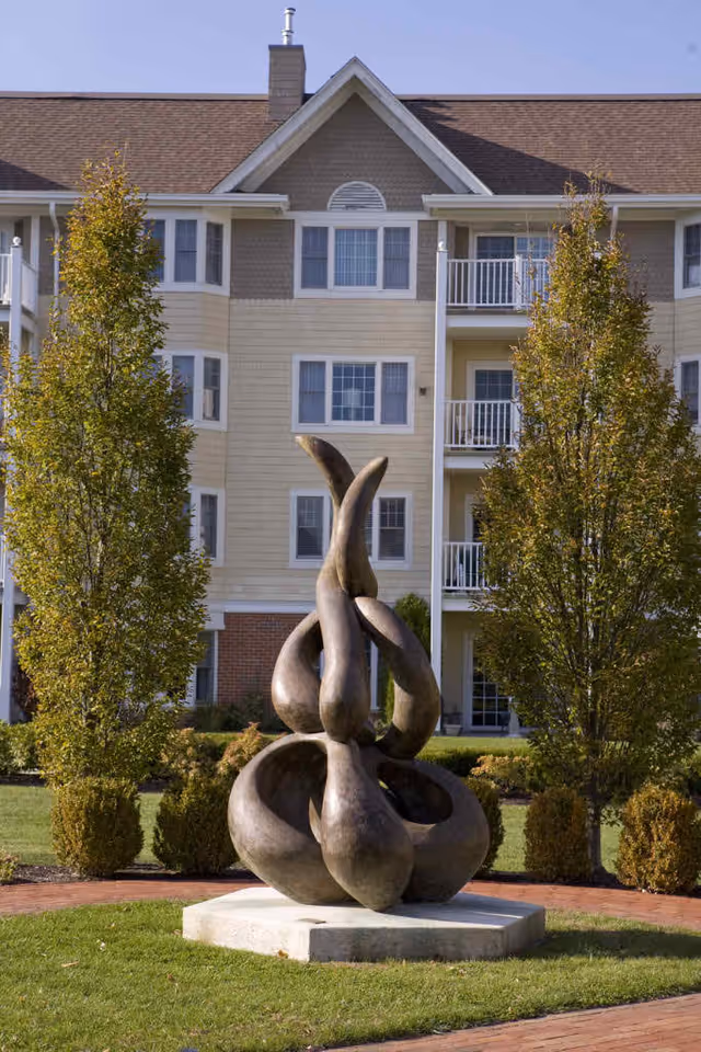Abstract bronze sculpture on a concrete base in a landscaped garden area with green grass and trimmed bushes, flanked by two tall trees. In the background, there is a multi-story residential building with balconies and multiple windows under a clear blue sky.