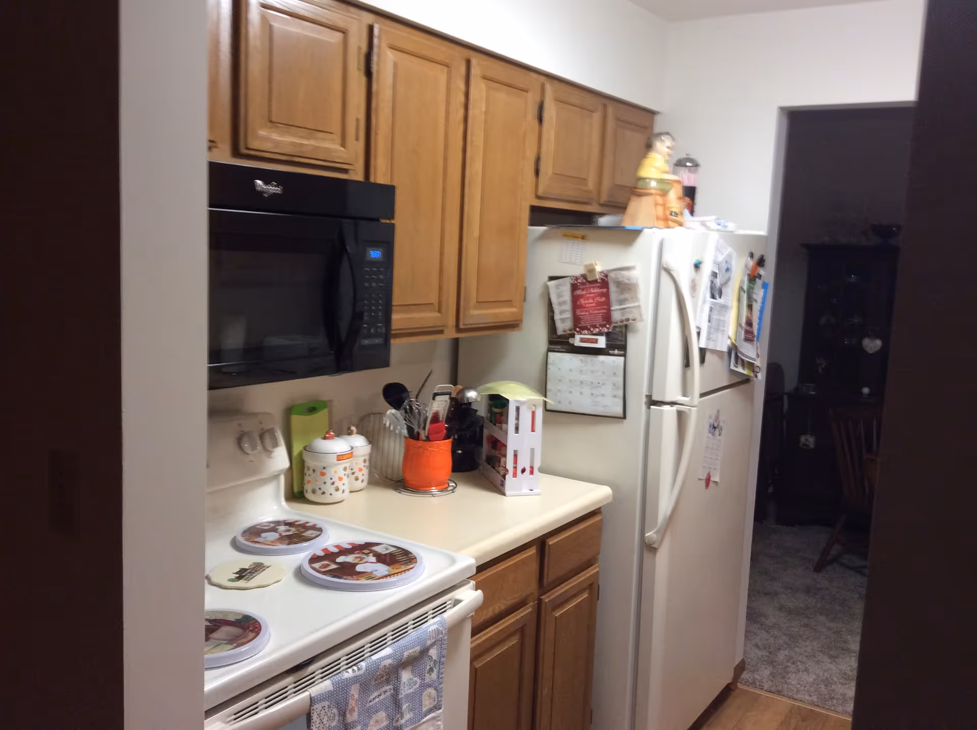 A small kitchen with wooden cabinets, a white refrigerator covered with papers and magnets, a white stove with decorative covers on the burners, a black microwave above the stove, and various kitchen utensils and containers on the countertop.