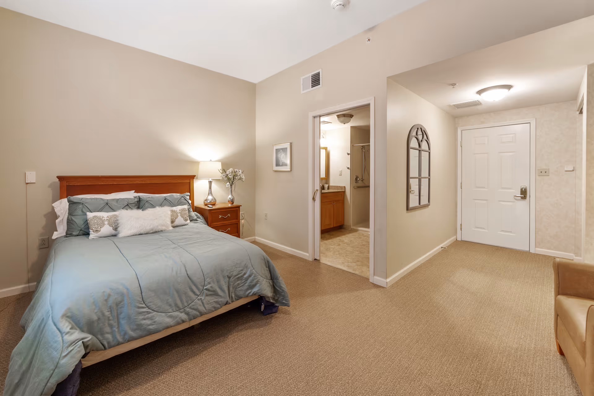 A bedroom with a neatly made bed featuring a blue comforter and decorative pillows. A wooden nightstand with a lamp and a vase of flowers is beside the bed. The room has beige walls and carpeted floor. There is an open doorway leading to a bathroom with a vanity and shower visible. Another doorway leads to a hallway with a white door and a wall-mounted mirror.