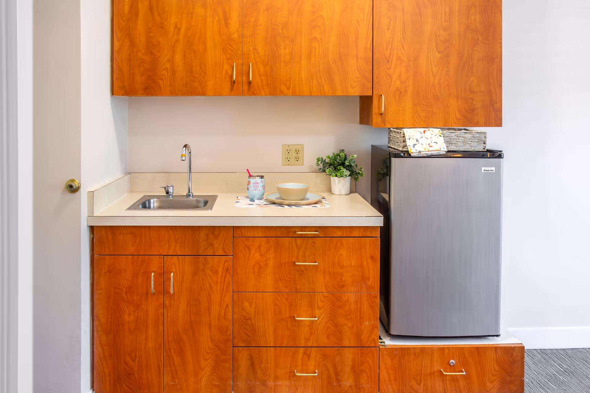 A small kitchenette area with wooden cabinets, a stainless steel sink with a faucet, a countertop holding a bowl, plate, cup with a straw, and a small potted plant. To the right, there is a compact stainless steel refrigerator with a woven basket and cloth on top.