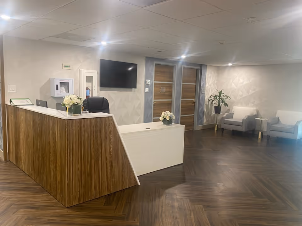 A modern reception lobby with a wooden front desk, seating area, wall-mounted TV, and potted plants.