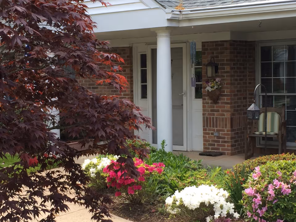Front entrance of a brick building with white columns, a porch with a chair, and a variety of colorful flowering bushes and plants in the garden area.