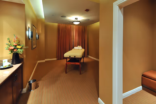 A softly lit massage room with a treatment table centered, warm-toned walls, a countertop with flowers to the left, and curtains at the back.