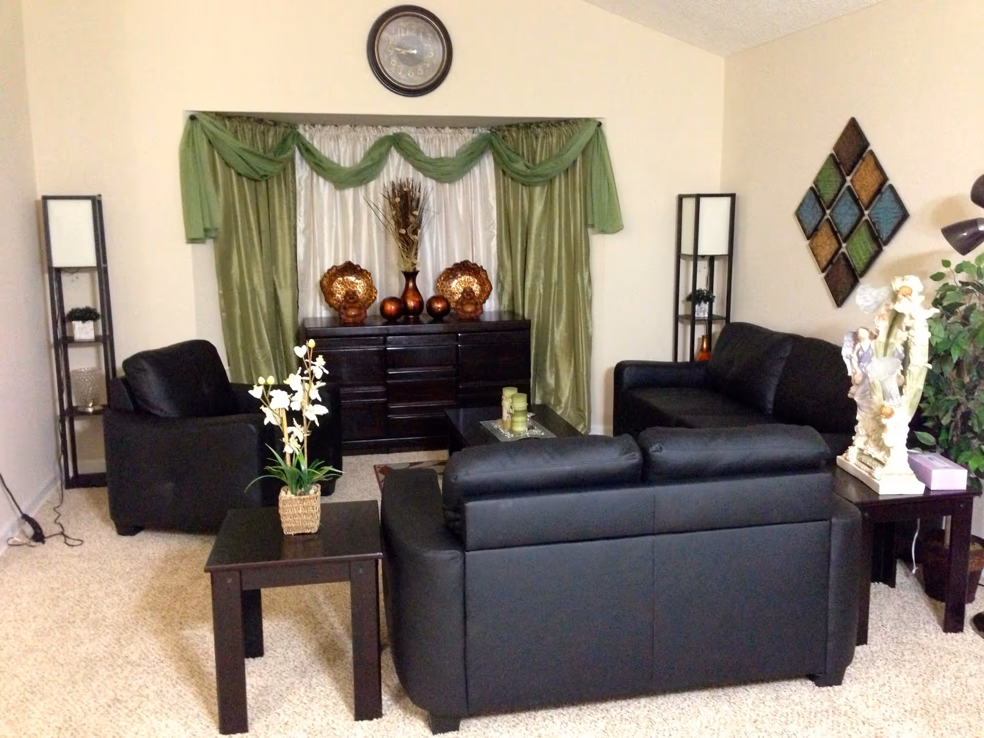 A cozy living room with black leather sofas and armchairs arranged around a dark wood coffee table. The room features green and white curtains with decorative vases on a dark wood sideboard against the back wall. There are two tall black shelving units with lamps on either side of the sideboard, a wall clock above the curtains, and a decorative wall art piece with diamond shapes on the right wall. A small table with a floral arrangement and another table with a decorative statue and a potted plant are also visible.