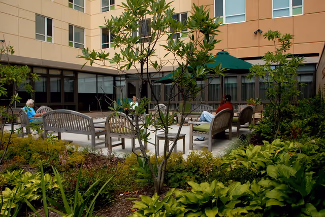 Outdoor courtyard area at Garito Manor At Union Square with wooden benches arranged around a central space. Several people are seated on the benches, surrounded by green plants and small trees. The building's beige exterior walls and windows form the backdrop, and a green patio umbrella is visible in the seating area.