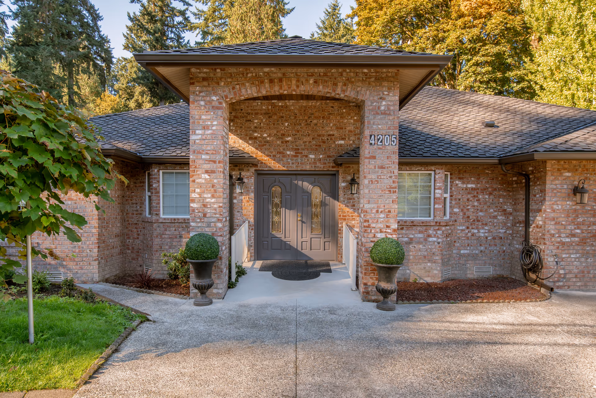 Front entrance of a brick building with a covered porch supported by a brick archway. The double front doors are dark-colored with decorative glass panels. Two potted topiary plants flank the entrance. The building number 4205 is displayed on the right side of the archway. Surrounding the building are trees and greenery.