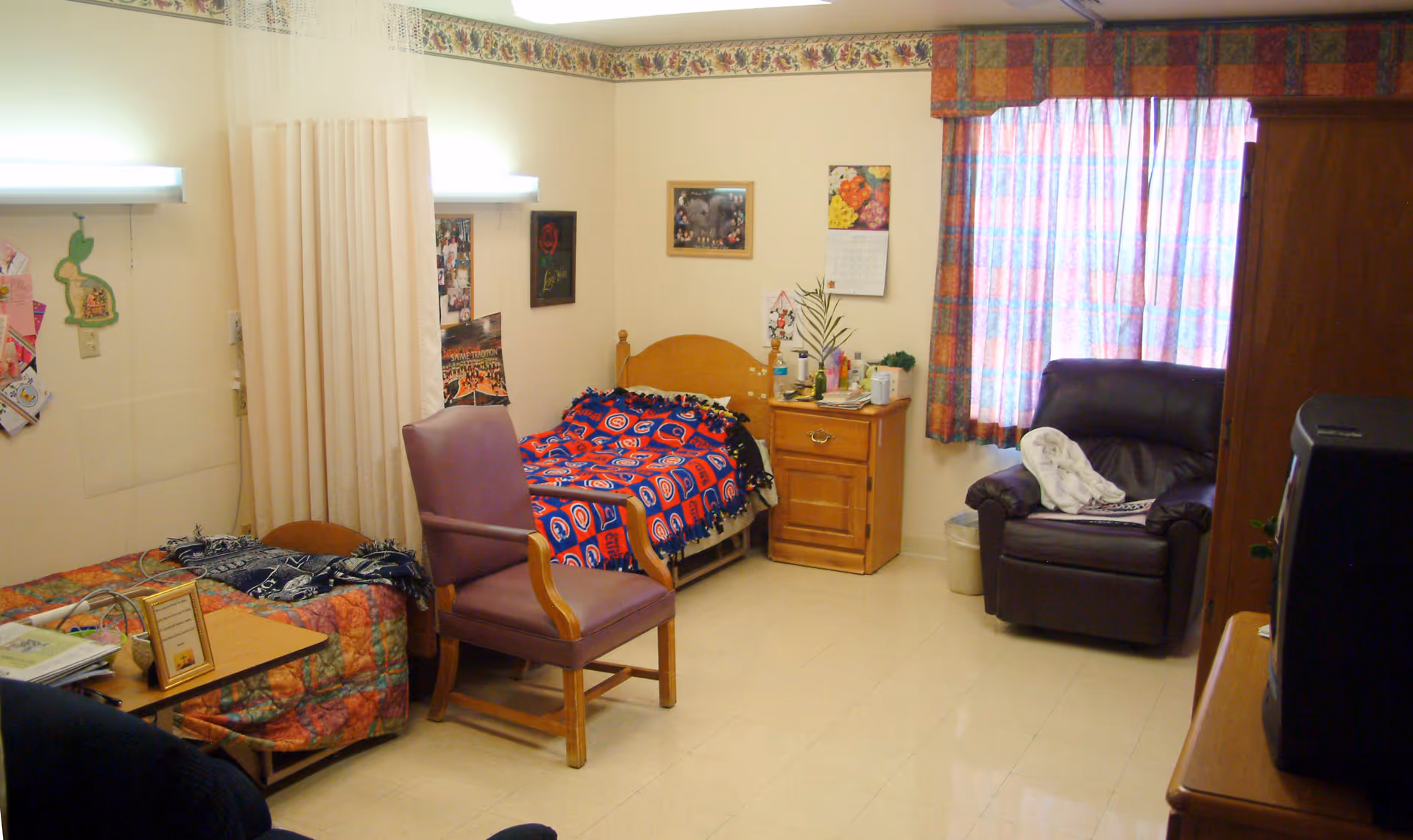 A small, cozy room with two single beds, each covered with colorful blankets. There is a wooden nightstand between the beds with various items on top. A purple armchair is placed near one bed, and a dark brown recliner chair is positioned near a window with patterned curtains. The walls have some pictures and a calendar hanging, and the floor is tiled.