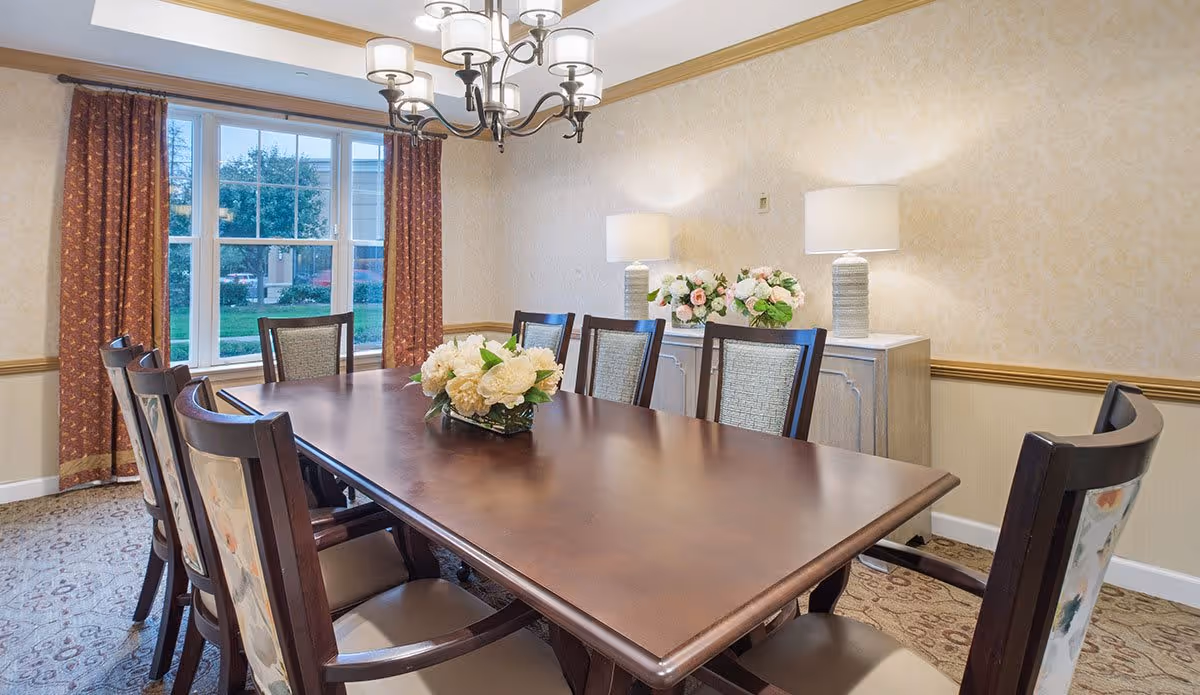 A dining room with a large wooden table surrounded by eight chairs, a floral centerpiece on the table, two lamps on a sideboard against the wall, and a window with red curtains showing greenery outside.