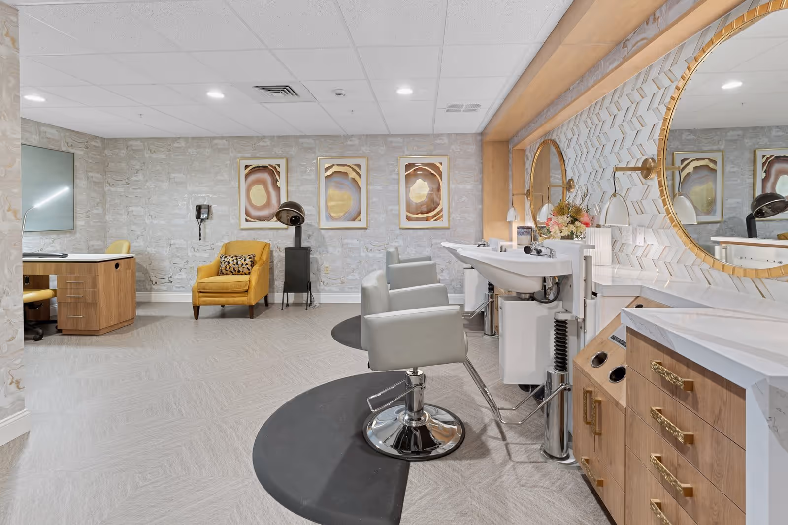 A stylish salon area in a senior living facility featuring two gray salon chairs in front of sinks and large round mirrors with gold frames. The room has light-colored walls with decorative artwork, a yellow armchair with a patterned pillow, a hair dryer, and a wooden desk with a chair. The floor is light gray with a subtle pattern, and the ceiling has recessed lighting.