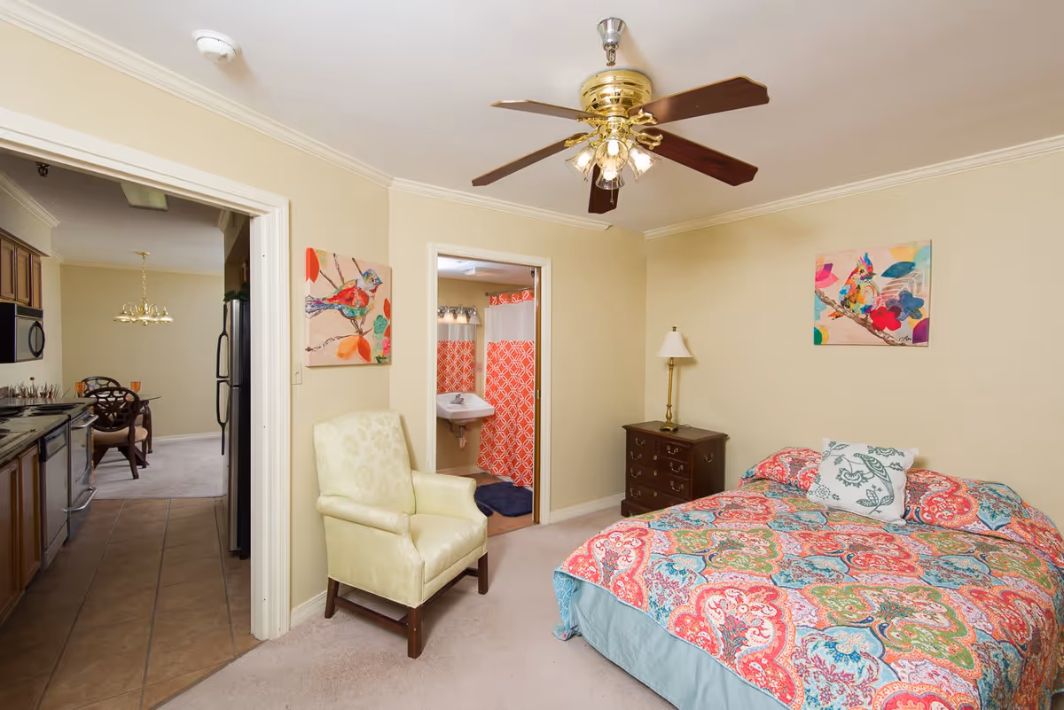 A cozy bedroom with a colorful patterned bedspread, a white armchair, a wooden nightstand with a lamp, and two colorful bird paintings on the walls. The room has beige walls and carpet. A ceiling fan with lights is mounted on the ceiling. The bedroom opens to a bathroom with a red and white shower curtain and a sink, and also connects to a kitchen area with wooden cabinets and a dining table visible in the background.