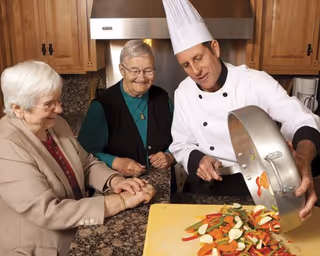 A chef wearing a white uniform and tall chef hat is pouring cooked mixed vegetables from a pan onto a cutting board while two elderly women watch and smile in a kitchen setting with wooden cabinets and granite countertops.