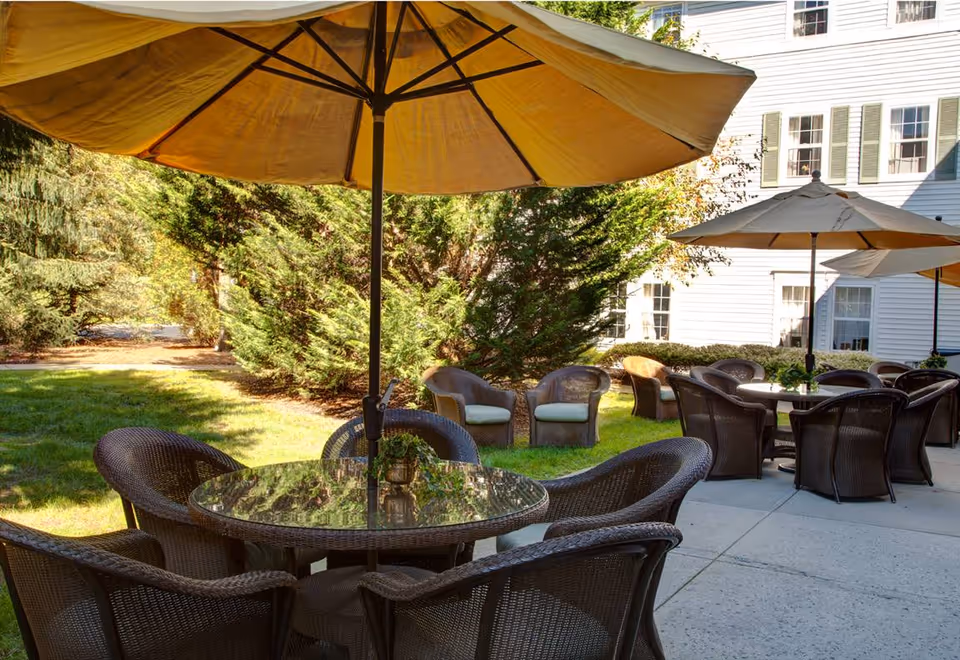 Outdoor patio area with round glass-top tables, wicker chairs and large umbrellas on a lawn beside a white building.