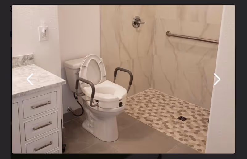 A bathroom with a white toilet equipped with safety rails on both sides, a marble countertop with drawers on the left, and a walk-in shower area with hexagonal patterned floor tiles and a grab bar on the wall.