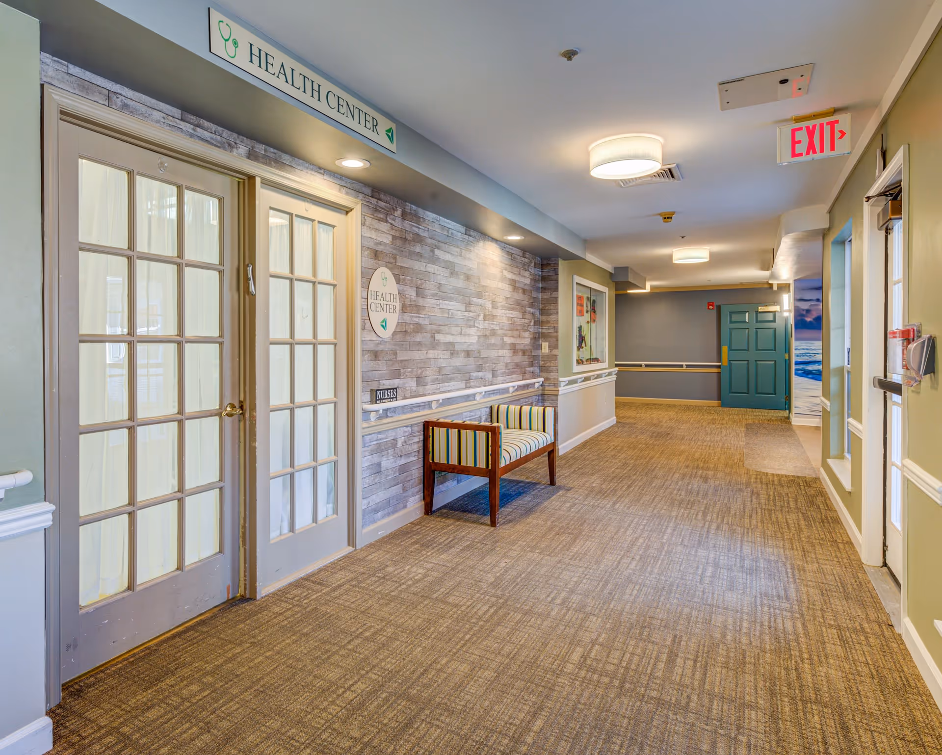 A well-lit hallway in a senior living facility with carpeted floors and light green walls. On the left side, there are double glass doors labeled 'Health Center' with a small striped bench nearby. The hallway has ceiling lights, handrails along the walls, an exit sign, and a teal door at the far end.