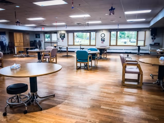 A spacious room with wooden flooring and several round tables surrounded by chairs, some with armrests and some without. The room has large windows letting in natural light and decorations hanging from the ceiling. There are stacked chairs and a door visible in the background.