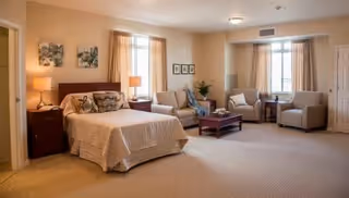 Spacious bedroom suite with a bed, nightstands, lamps, and a seating area of chairs and a coffee table by windows.