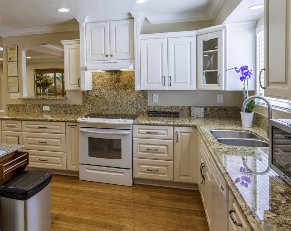 A bright and clean kitchen with white cabinets, granite countertops, a white stove, a double sink, a microwave, and a stainless steel trash can. There is a purple orchid plant on the countertop near the window, and a vase with yellow flowers is visible through a pass-through window to an adjacent room.