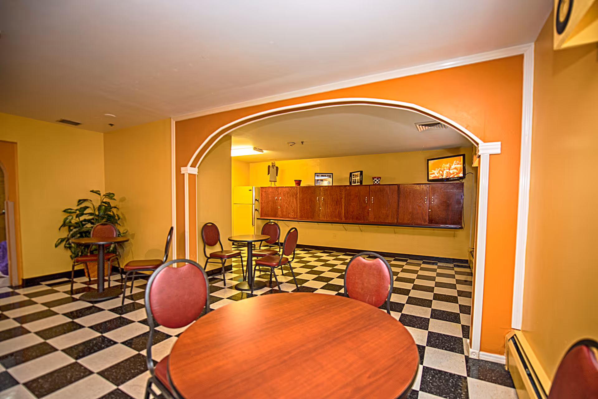 Interior view of a common area with checkered black and white flooring, round wooden tables with red cushioned chairs, a potted plant in the corner, and a yellow wall with wooden cabinets and a small TV mounted above them.
