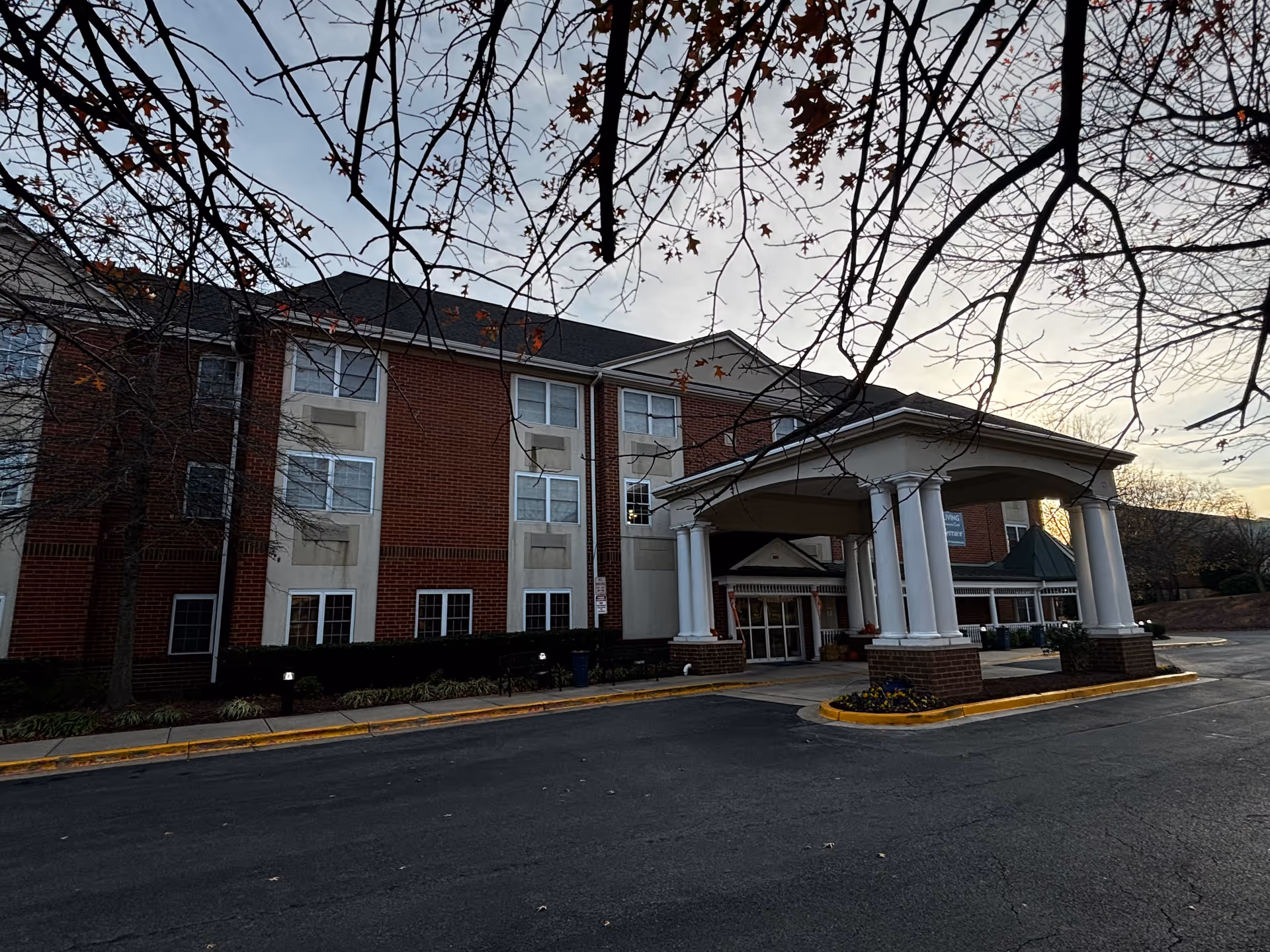 Front exterior of a three-story brick senior living building with a covered entrance supported by white columns and bare tree branches overhead.