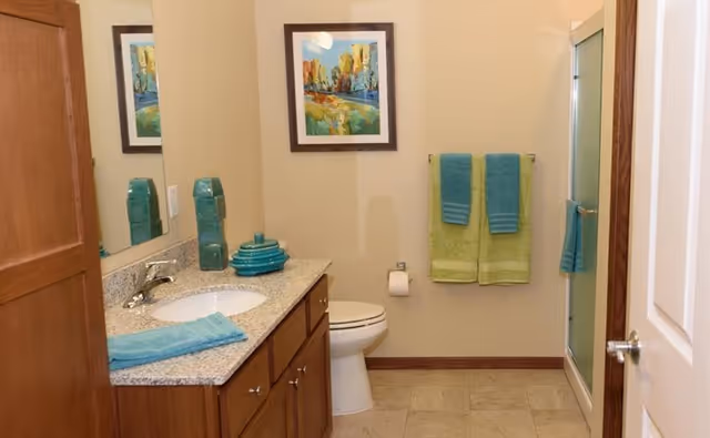 A clean bathroom with a wooden vanity topped with a granite countertop, a sink, and teal decorative items. There is a toilet next to the vanity, a towel rack with green and teal towels, a framed colorful landscape painting on the wall, and a shower with a frosted glass door.