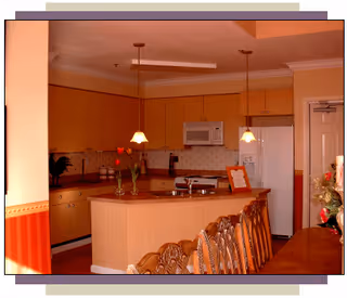 Open kitchen with an island, pendant lights, wooden cabinets, a refrigerator and microwave, and dining chairs in the foreground.