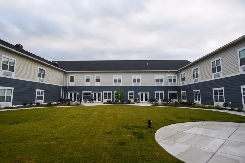 Outdoor courtyard area of Traditions at Hunter Station with a large green lawn surrounded by a two-story building with beige and blue siding, multiple windows, and several patio seating areas under a cloudy sky.