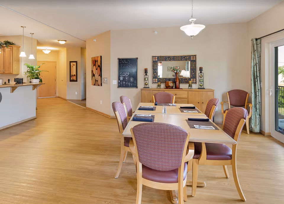 Bright communal dining room with a table and several chairs, a buffet cabinet and mirror, and a view into the kitchen area.
