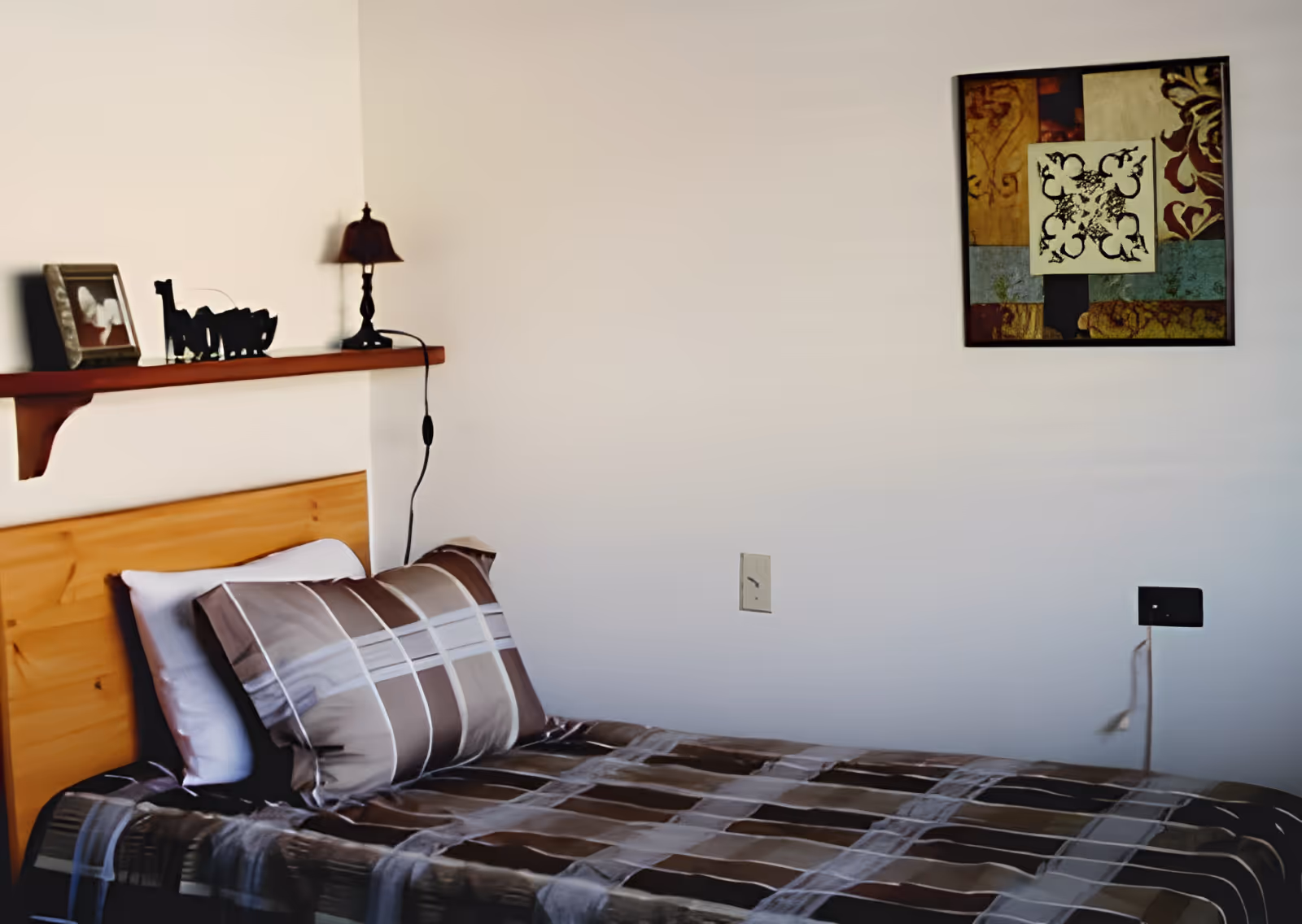 A single bed with a wooden headboard, covered with a brown and beige plaid bedspread and matching pillows. Above the bed is a wooden shelf holding a small lamp, a framed photo, and a decorative item. On the wall to the right of the bed is a colorful abstract painting.