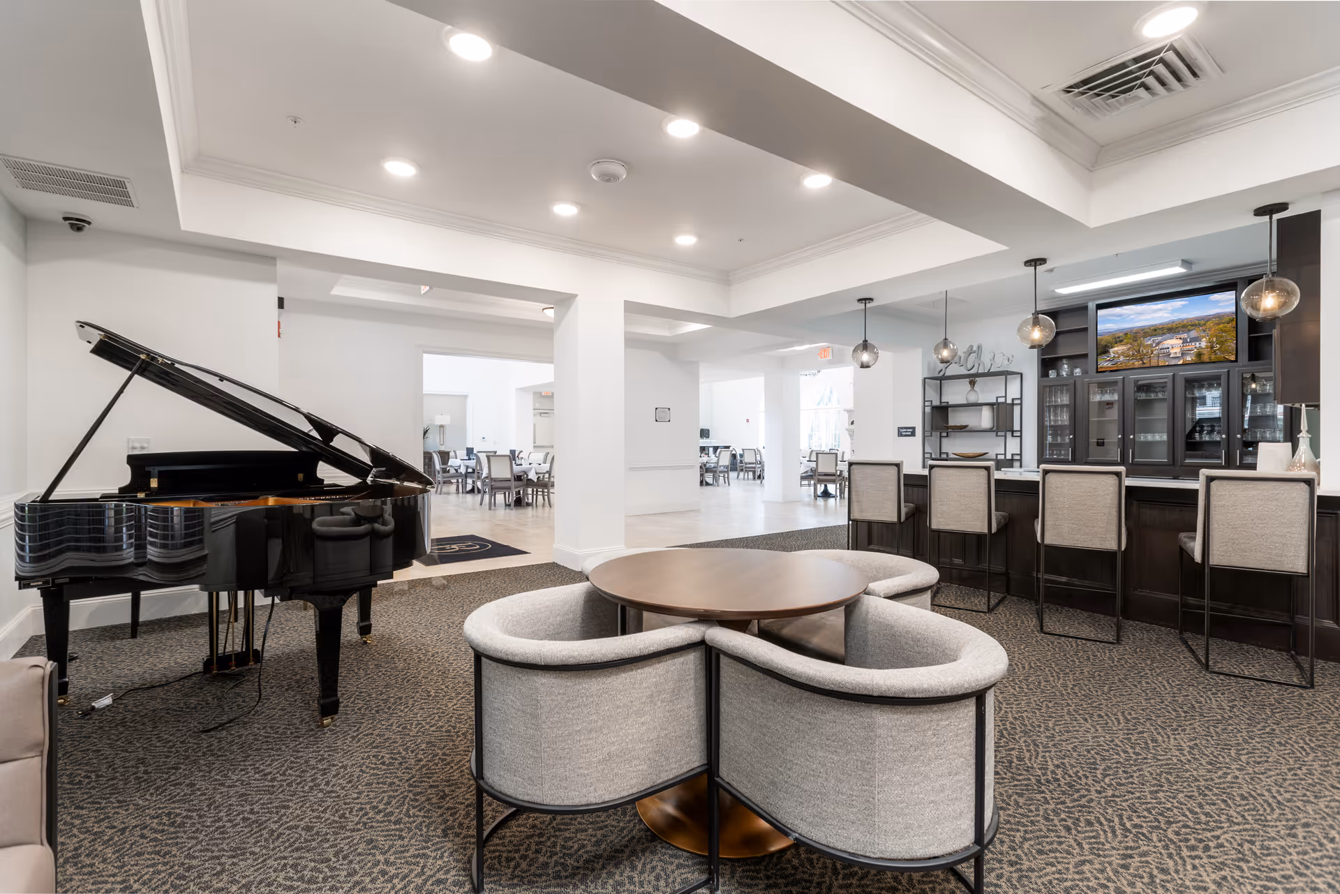Bright communal lounge with a grand piano, circular seating around a round table, and a bar with stools and TV.
