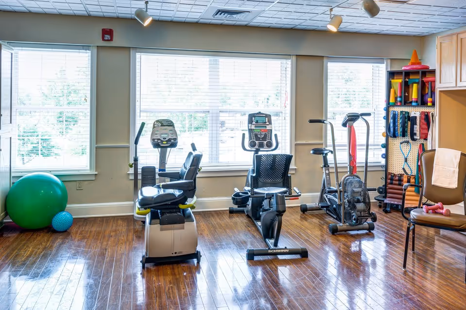 Bright exercise room with stationary bikes, an air bike, exercise balls, and shelves of fitness equipment in front of large windows.