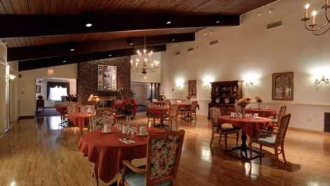 Spacious dining room with round tables, red tablecloths, and chandeliers in a senior facility.