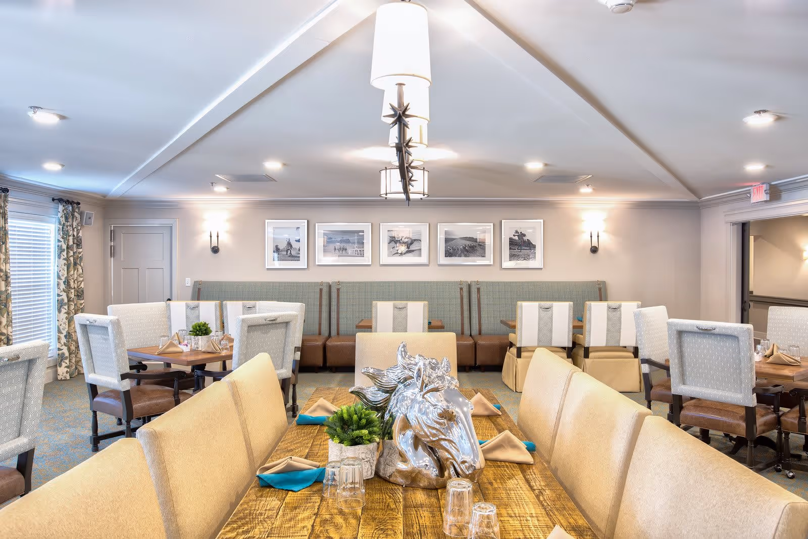A bright and elegant dining room with multiple tables and cushioned chairs. The tables are set with napkins, glasses, and small decorative plants. A large wooden table in the foreground features a silver horse head sculpture as a centerpiece. The walls are adorned with framed black and white photographs, and the room is softly lit with ceiling lights and wall sconces.