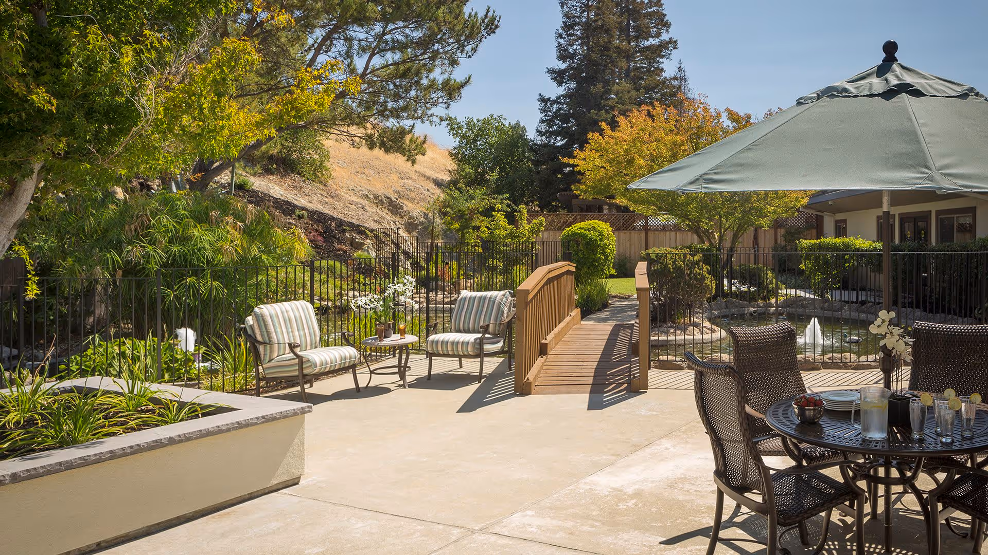 Outdoor patio area with cushioned chairs and a small table on the left, a wooden bridge over a small pond with a fountain in the center, and a round table with chairs and a large green umbrella on the right. The area is surrounded by trees, shrubs, and a fence, with a building visible in the background under a clear blue sky.
