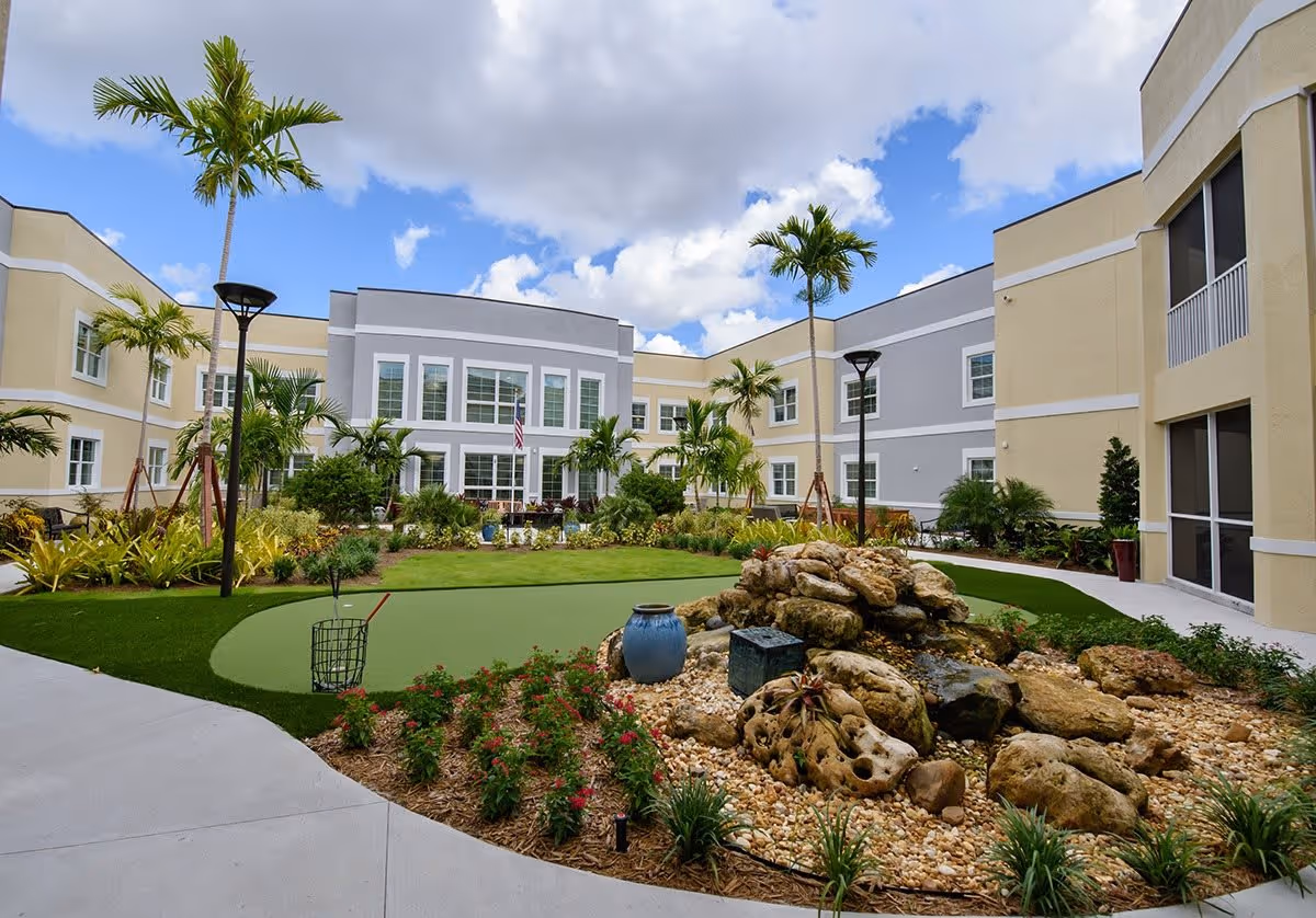 Courtyard of a senior living community with a landscaped putting green, rock fountain, palm trees, and a surrounding two-story building.