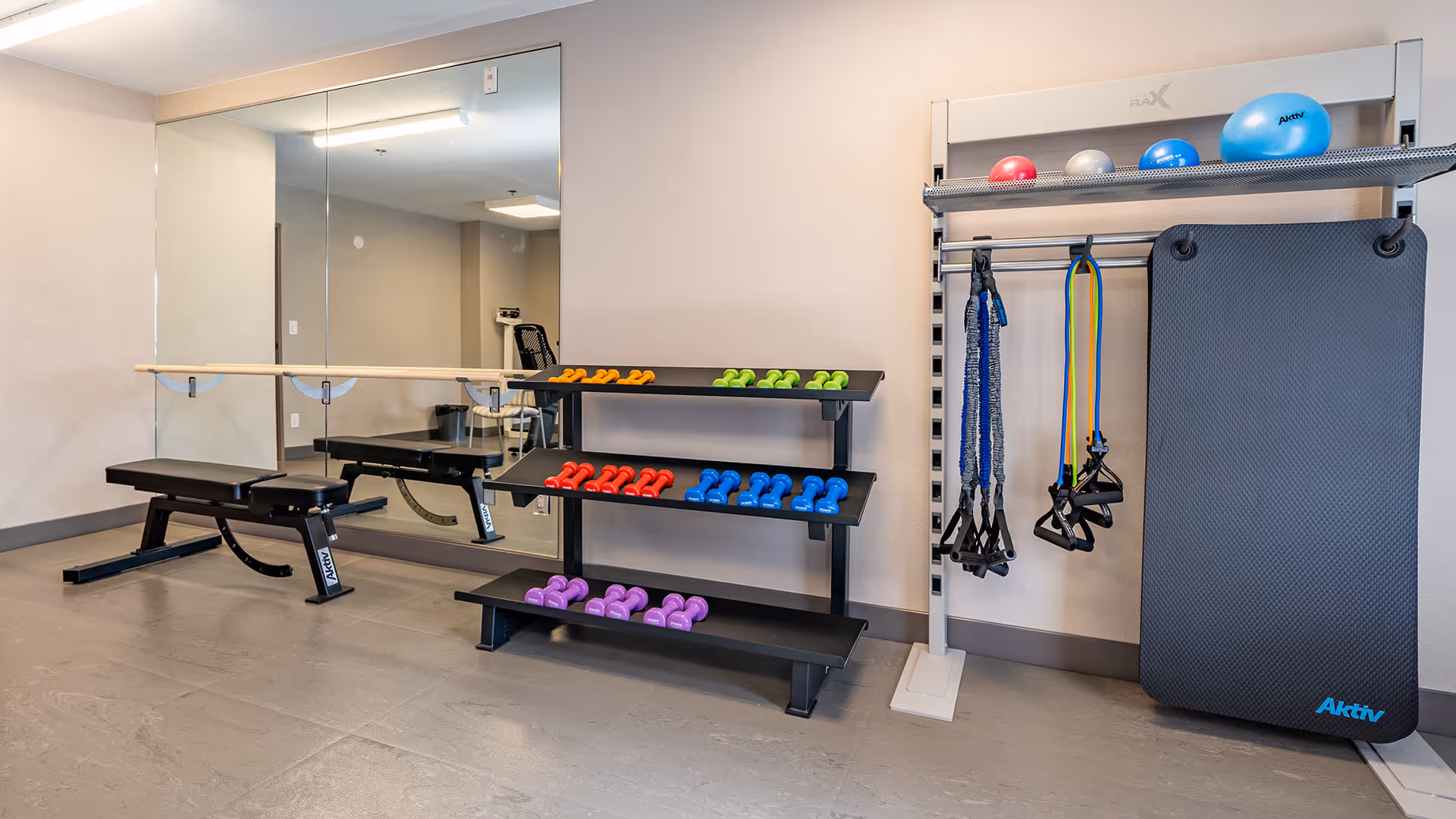 A small fitness room with a bench, a rack of colorful dumbbells, resistance bands hanging on a rack, exercise balls on the top shelf, and a large exercise mat leaning against the wall. There is a large mirror on the wall behind the bench and dumbbells.