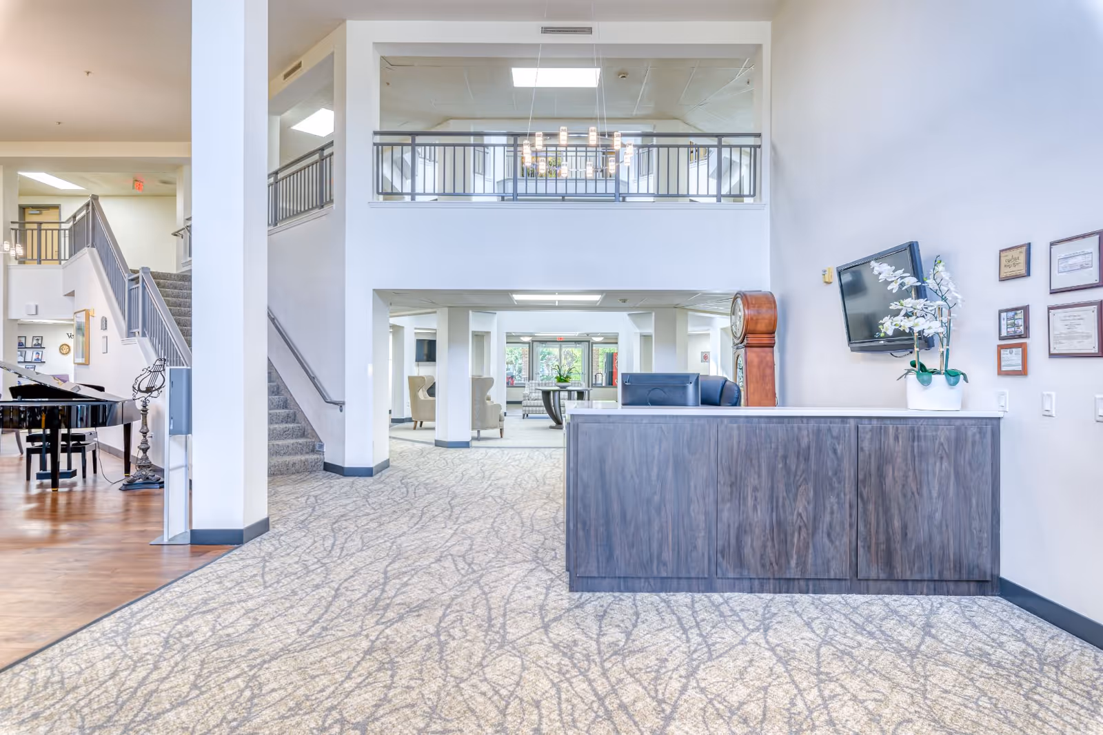 Bright two-story senior living lobby with a reception desk, staircase, piano, and seating area.