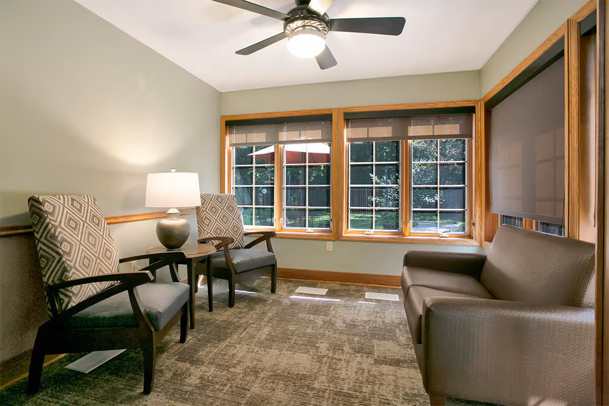 A cozy sitting area with two patterned armchairs and a small table with a lamp between them, a brown sofa, large windows with wooden frames showing a view of a green outdoor space, and a ceiling fan with a light.