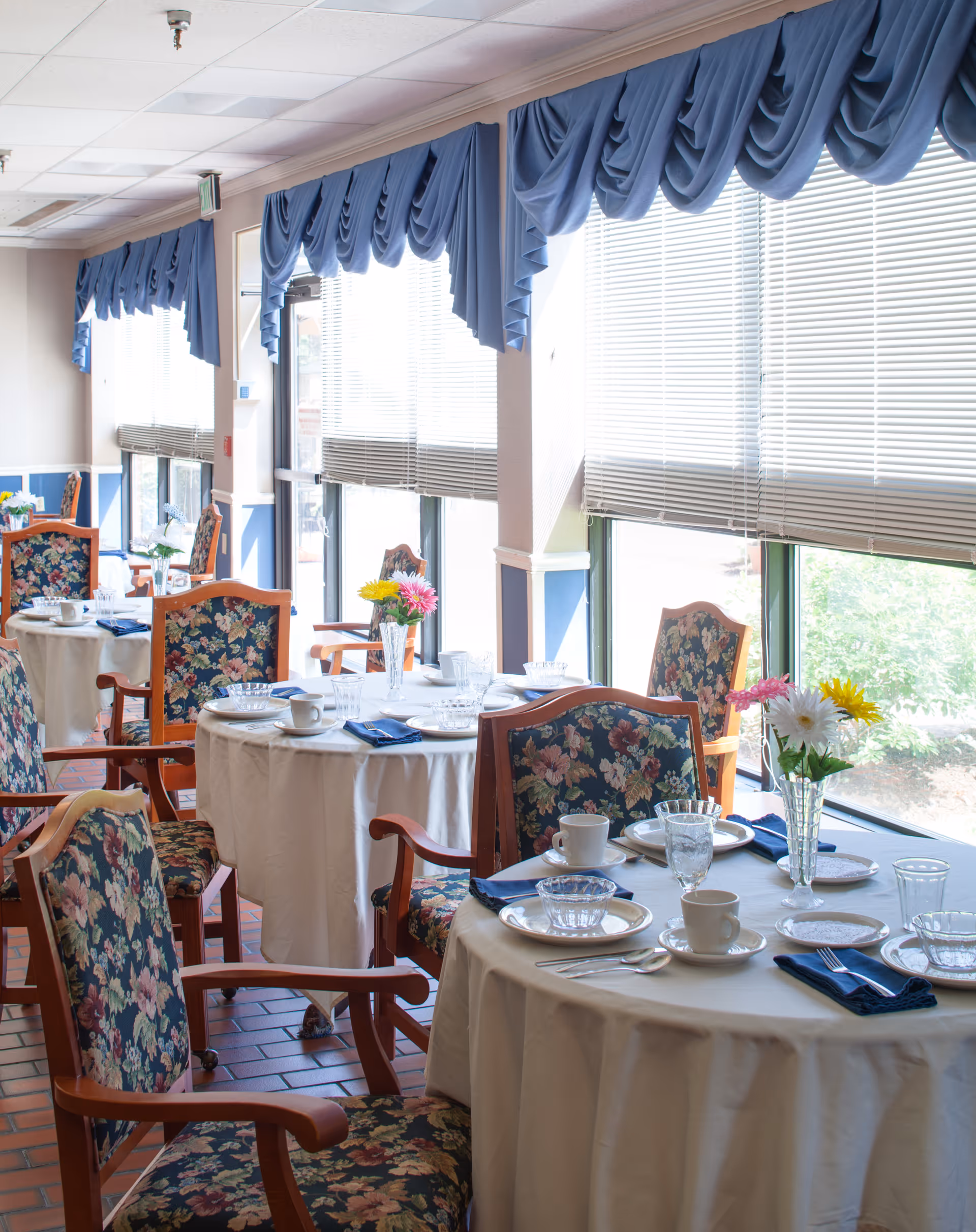 Dining room with round tables set for meals, floral-upholstered chairs, vases of flowers, and large windows with blue valances.